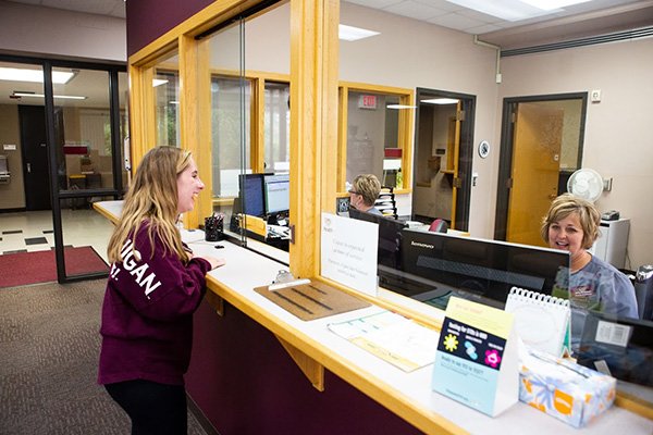CMU-Health-Services-600x400 A student in maroon shirt interacting with a woman at a desk and computer.