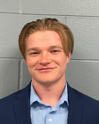 A man with blonde hair wearing a navy suit coat and blue button up smiles.