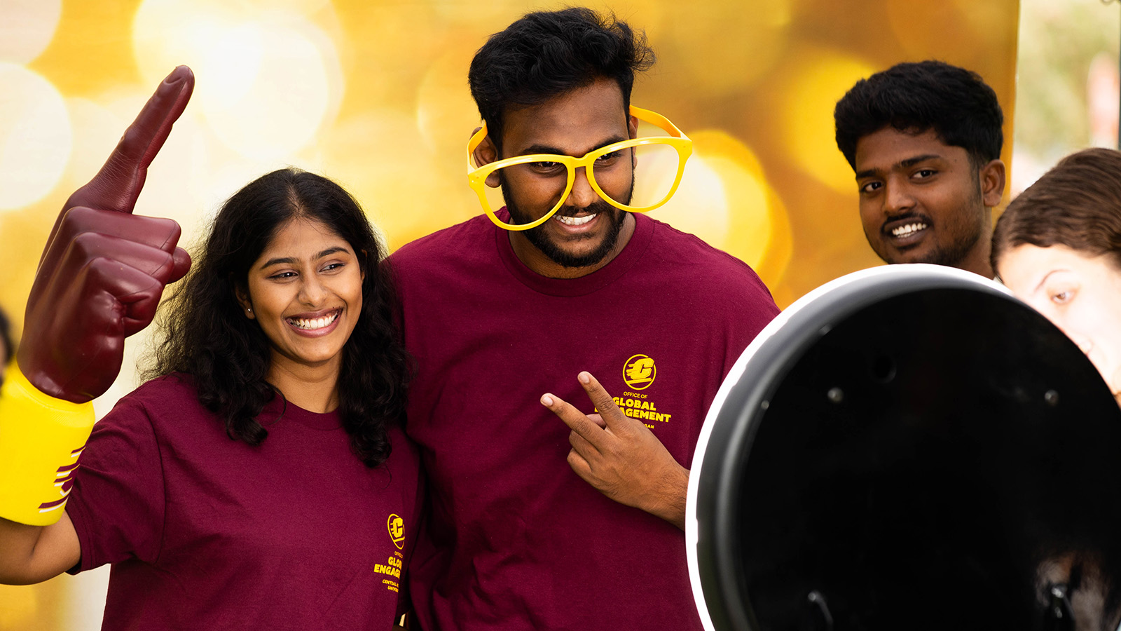 Two students smile and pose against a gold backdrop for a photo. One student is wearing oversized glasses and another is wearing a foam hand.