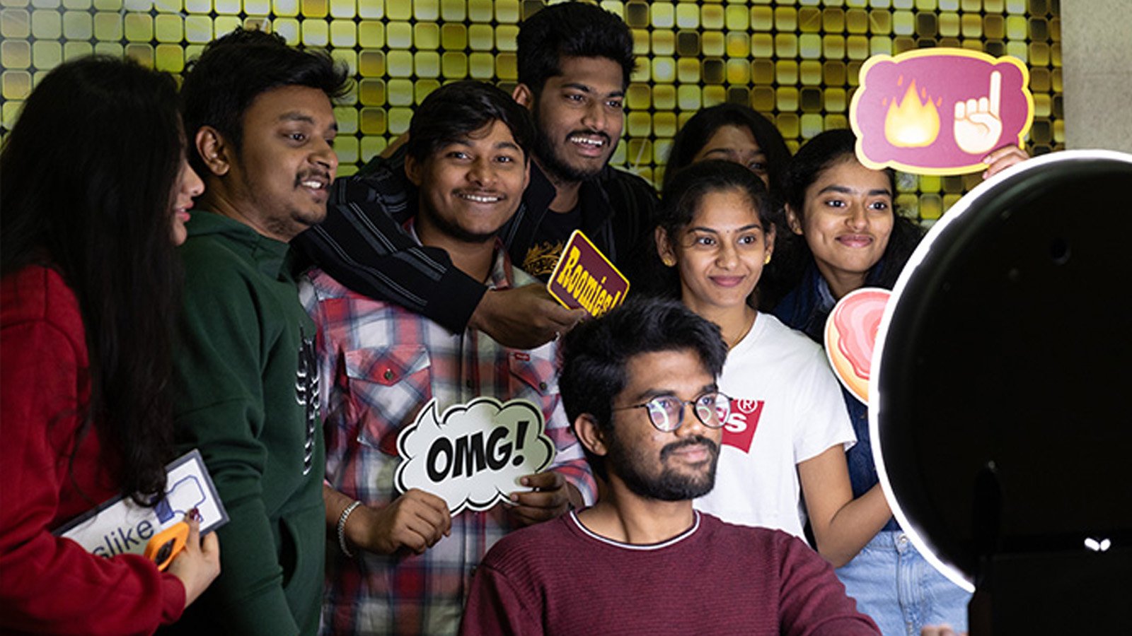 A group of students smile for a selfie-style photo in front of a camera and ring light