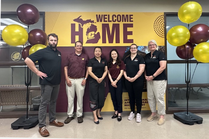 Six people stand in a line in front of a maroon and gold backdrop that says Welcome Home, with the state of Michigans outline replacing the O, and smile for a group picture in an office building. Maroon and gold balloons decorate the sides