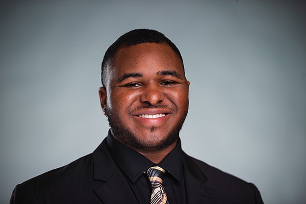 Professional headshot of a smiling Donovan Pitts-Reed wearing a black jackets, black shirt, and a plaid tie against a light grey background.