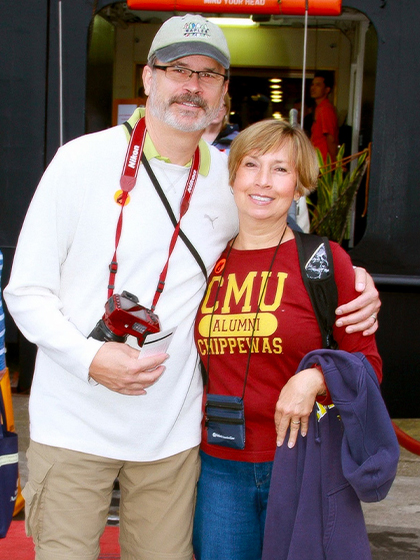 A man in a long sleeve white shirt and cargo khaki pants poses alongside a woman wearing a CMU Chippewas Alumni shirt and blue jeans.