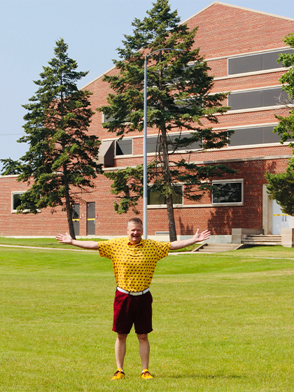 A man wearing a yellow polo and maroon shorts stands on grass in front of a building with his arms spread out.