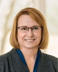 Sarah Opperman wears a grey sweater and blue top while smiling for the camera.