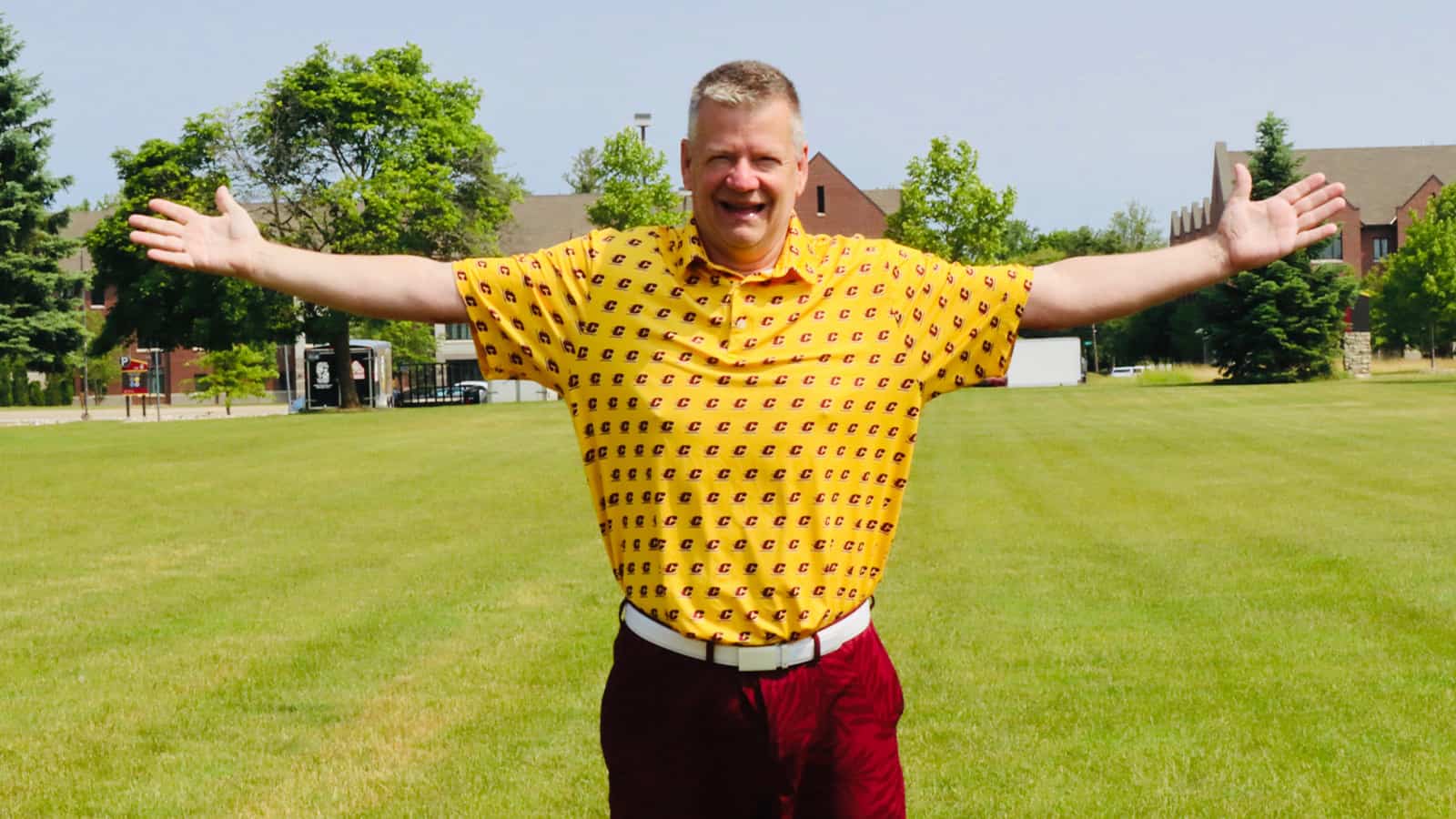 A man wearing a yellow polo and maroon shorts stands with his arms spread out.