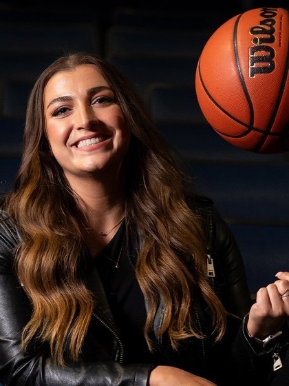 A woman in a black leather jacket smiles and tosses a basketball in the air while sitting.