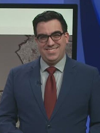 A man with dark hair wearing a suit and tie stands in front of a weather map on a TV screen.