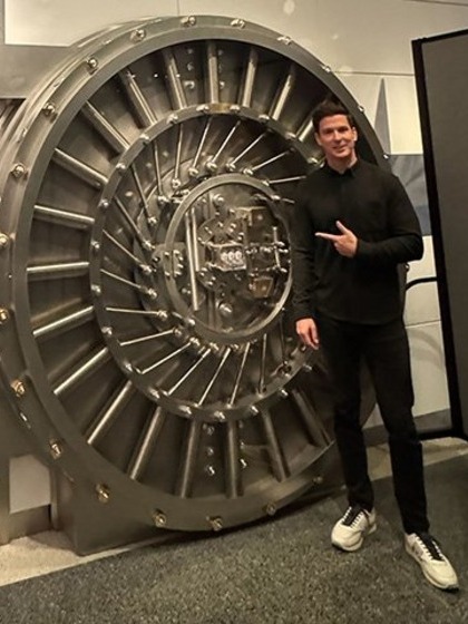 A man in sneakers and a dark track suit stands next to and points to an open bank vault leading to a museum exhibit.
