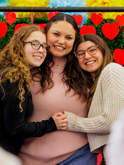 Close-up of three cheerful young women embracing and smiling in front of a backdrop decorated with red hearts and colorful tissue paper flowers.
