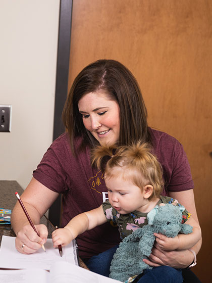 A woman sits at a desk, holding a baby while studying.