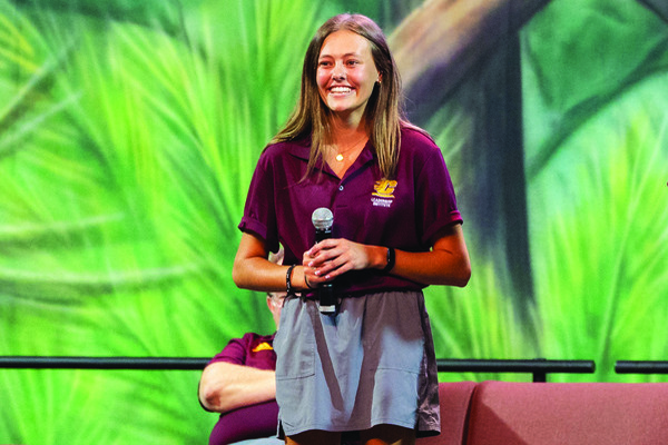 A woman wearing a maroon polo with a CMU logo, speaking on stage in front of a green background.