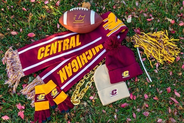 A collection of Central Michigan University spirit items laid out on the grass, including a football, a maroon and gold CMU scarf, a maroon beanie, winter gloves, a gold chain, and pom-poms.