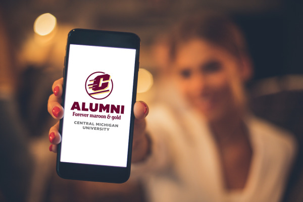 A woman holds a smartphone showing a white screen with the maroon and gold CMU Alumni logo and text showing Alumni Forever maroon & gold with Central Michigan University at the bottom.