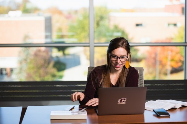 A person wearing glasses and a maroon shirt sits at a desk with a laptop displaying the CMU Action C logo, in front of a large window with an outdoor view.