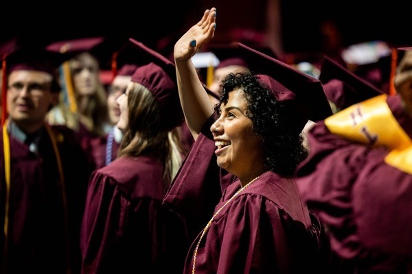 A CMU graduate with curly dark hair, wearing a maroon cap and gown, waves during a commencement ceremony.