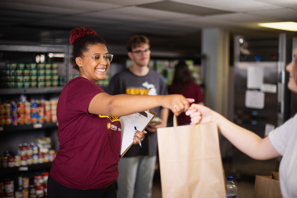 A student volunteer with glasses and a maroon t-shirt hands a brown paper bag to an outstretched hand at the CMU Food Pantry.