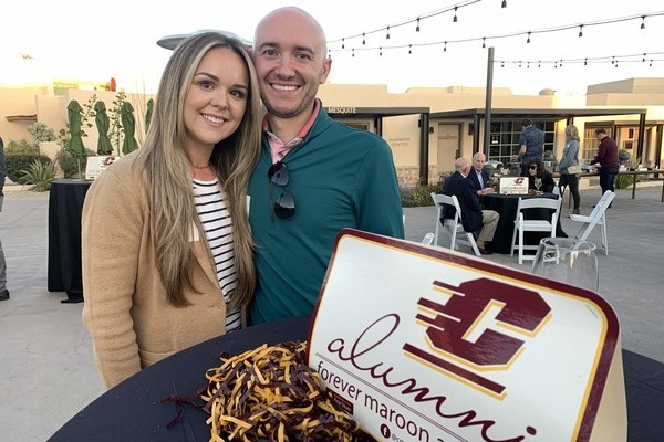 A couple pose behind a table with maroon and gold CMU alumni branding at an outdoor gathering.
