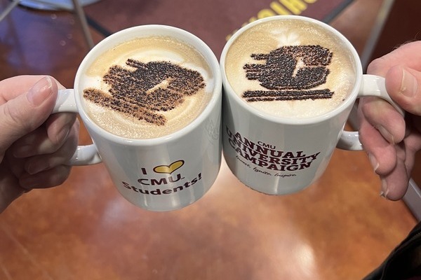 Two white mugs of lattes held by hands, each featuring latte art depicting the CMU Action C.