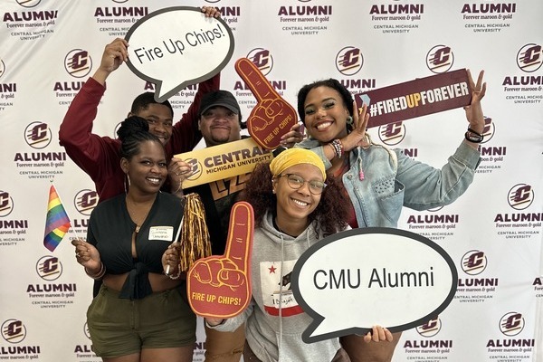 A group of five Central Michigan University alumni pose in front of a CMU alumni backdrop, holding spirit items including foam fingers, speech bubbles and signs.