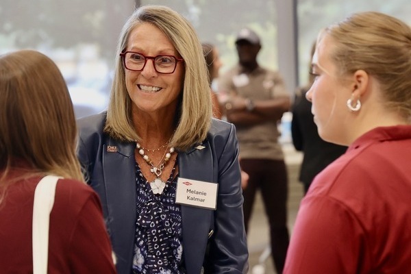 A woman with blonde hair and glasses, wearing a name tag, engages in conversation with two other people at an indoor event.