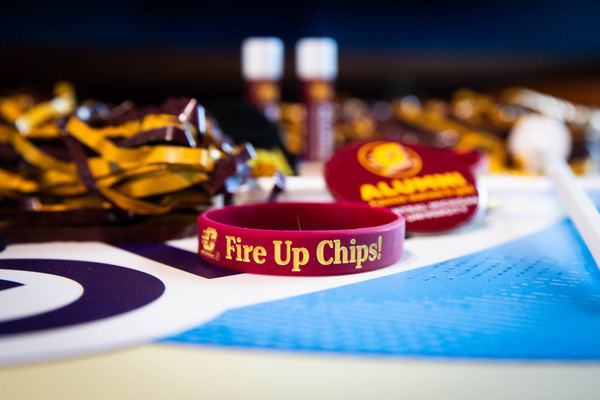A close-up of Central Michigan University spirit items on a table, including a maroon 'Fire Up Chips!' wristband and a maroon alumni button.