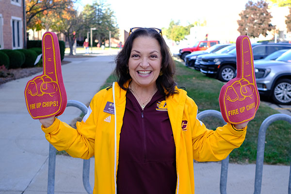 A smiling woman, wearing maroon and gold Central Michigan University apparel, holding up two maroon foam fingers that say "Fire Up Chips!"