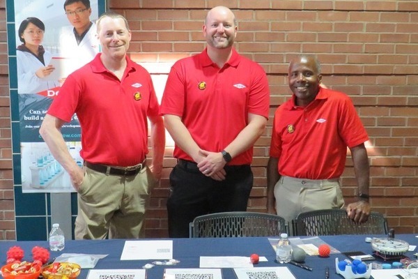 Three men in red polo shirts stand behind a table covered with a blue tablecloth, in front of a brick wall.