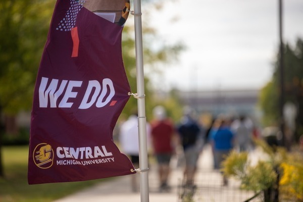 A maroon Central Michigan University flag featuring the text 'We Do.' flies near a campus walkway with students walking in the background.