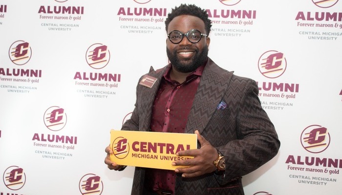 A CMU alum with glasses and a beard holds a gold sign with the CMU logo, against a backdrop featuring CMU Alumni branding.