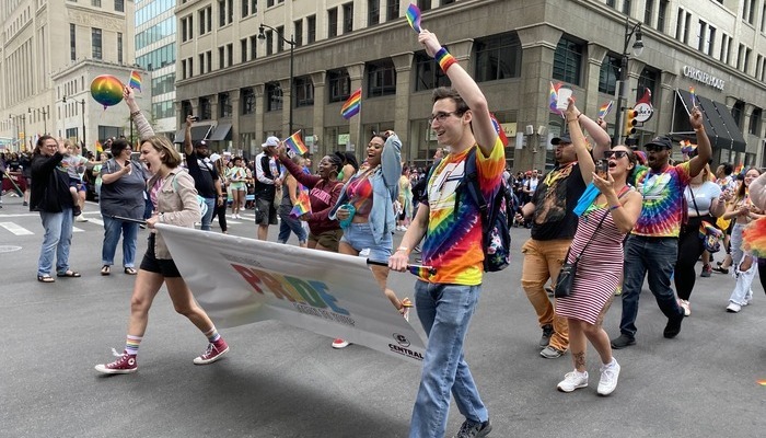 Members of CMU's LGBTQIA+ community walk in parade while holding a pride sign and flags.