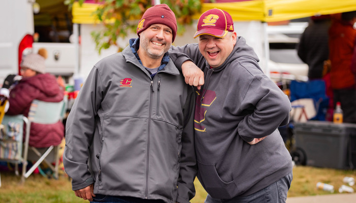 Two alumni in CMU gear pose in front of a tailgate tent at a football game.