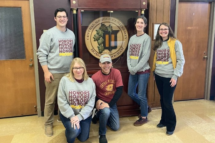 Five individuals pose for a photo in front of the Central Michigan University Honors Program display. They are wearing matching gray crewneck sweatshirts.
