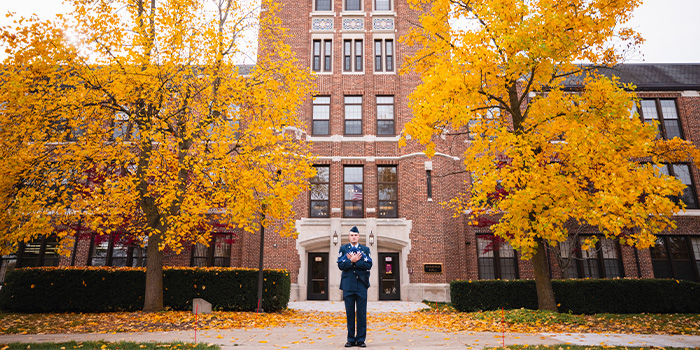 A person in a military uniform stands on a sidewalk in front of a building.