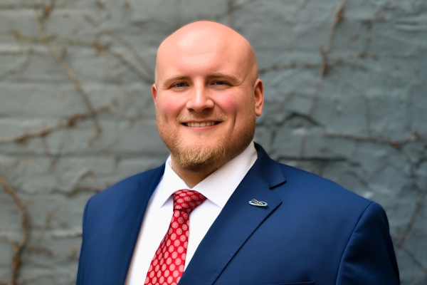 A man with a trimmed beard is wearing a blue suit jacket, white shirt, and red patterned tie. He is standing in front of a textured gray wall with dried vines.