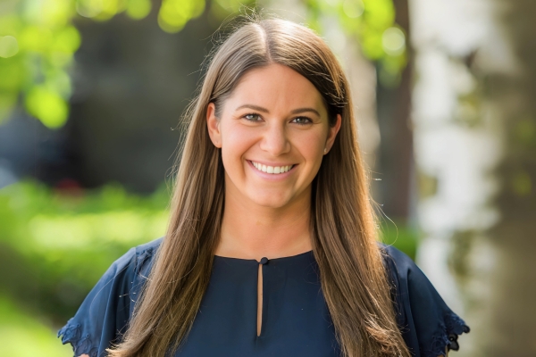A woman with long straight brown hair is wearing a navy blouse and standing outdoors. She is smiling, with greenery and sunlight visible in the blurred background.
