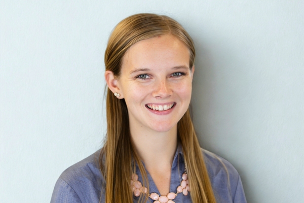 A young woman with long straight hair smiles at the camera. She is wearing a blue button-up shirt and a pink necklace, posed in front of a plain light background.
