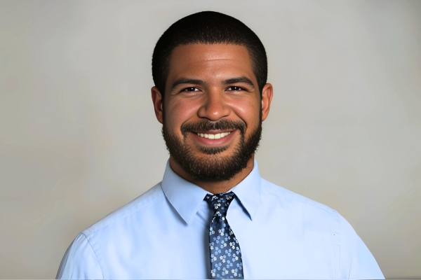 A man with short dark hair and a full beard is smiling at the camera. He is wearing a light blue dress shirt and a blue patterned tie. The background is a plain, neutral color.