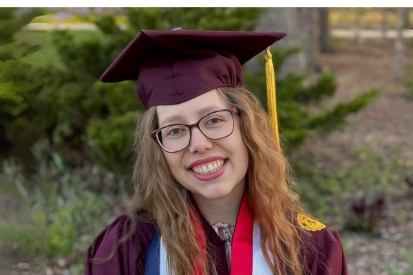 A young woman wearing a maroon graduation cap and gown smiles outdoors. She has long wavy hair, glasses, and wears honor cords and a medal around her neck.