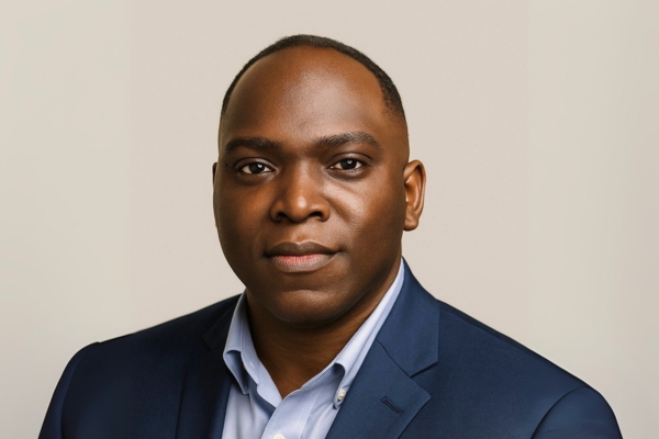 A smiling man in a light brown plaid suit and colorful bow tie poses in front of a light-colored curtain backdrop.