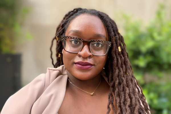 A woman with tortoiseshell glasses looks at the camera. She is wearing a light-colored top, a small gold necklace, and dark lipstick. Green foliage is blurred in the background.