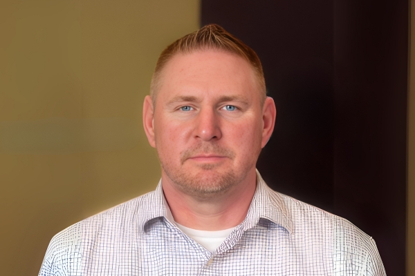 A man with short blond hair and a trimmed beard wears a light plaid button-up shirt. He poses against a neutral indoor background with soft lighting.