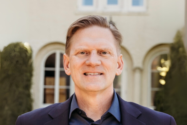 A man with short light brown hair is wearing a dark blazer over a collared shirt. He is standing in front of a light-colored building with arched windows and trimmed bushes.