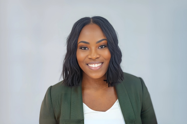 A woman with shoulder-length wavy black hair is wearing a dark green blazer over a white top. She is posed against a plain light-colored background and is smiling at the camera.