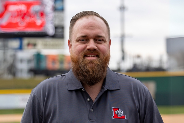 A man with a full beard is wearing a gray polo shirt with a red and white "L" logo. He is standing on a baseball field with a scoreboard and stadium structures in the blurred background.