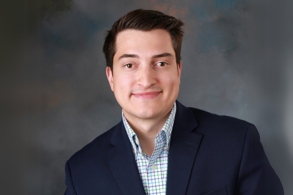 A young man with short dark hair is smiling slightly, wearing a navy blazer over a checkered dress shirt. He is posed in front of a neutral studio background.