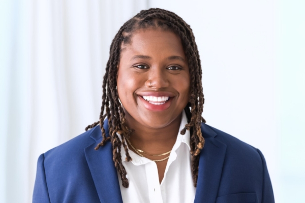 A smiling person is wearing a blue blazer over a white shirt and layered gold necklaces. The background is bright with soft white curtains.
