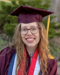 A young woman wearing a maroon graduation cap and gown smiles outdoors. She has long wavy hair, glasses, and wears honor cords and a medal around her neck.