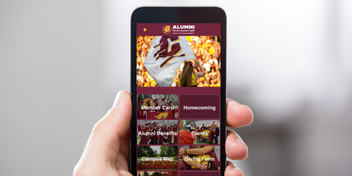 A hand holds a smartphone displaying the homescreen of the CMU Alumni App, featuring a banner image and menu options like Member Card, Homecoming, Alumni Benefits, Events, Campus Map and Giving Form.