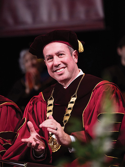 A man in a maroon robe wears a hat and neck medallion at a college commencement ceremony.